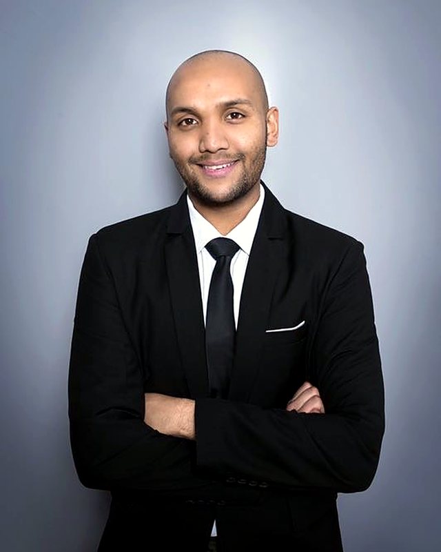 Portrait of Craig Cameron in a dark suit, smiling with his arms folded